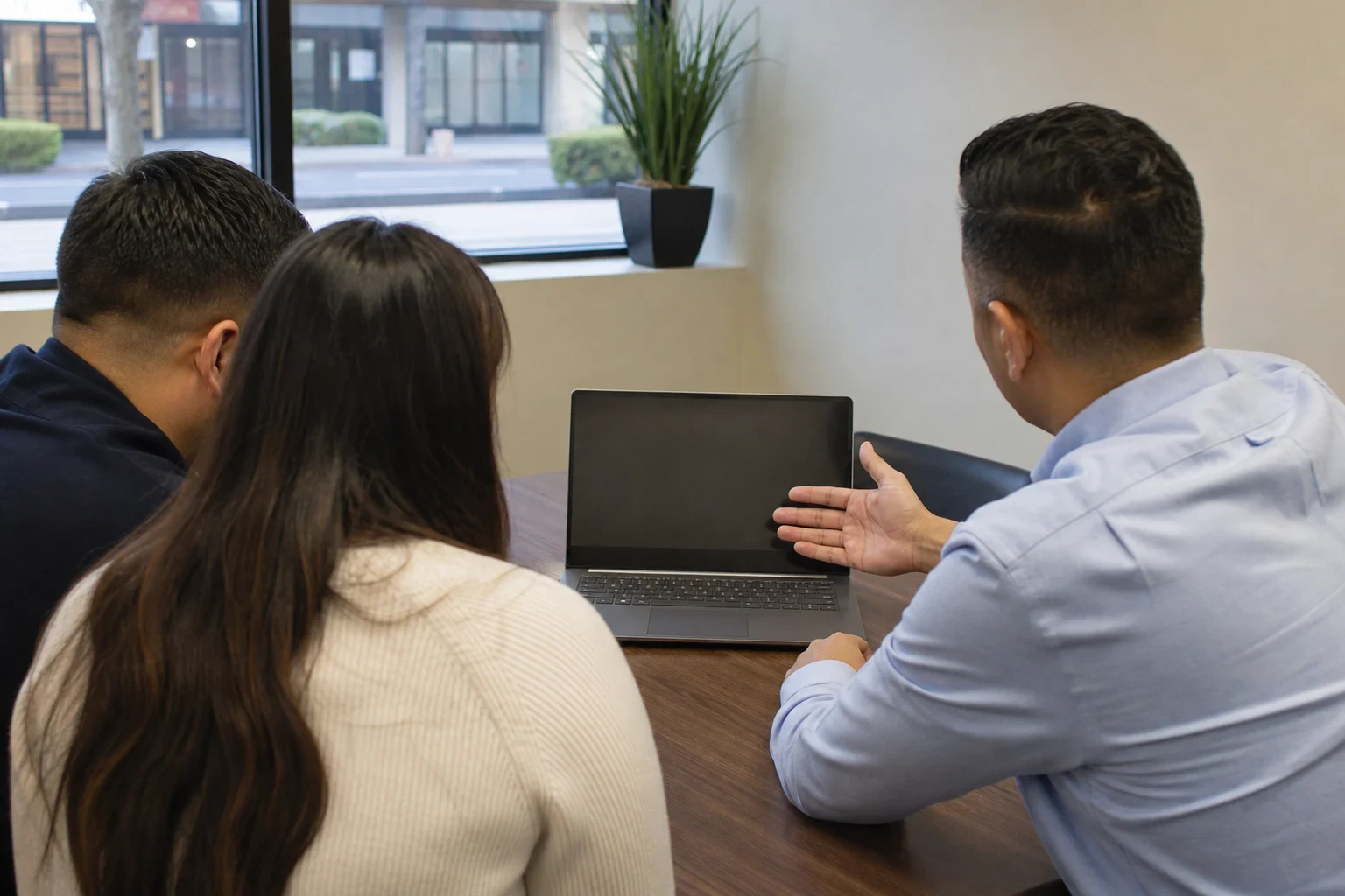 Agent in a light blue shirt gestures toward a laptop on a wood desk while a couple sits beside him, near a window with a potted plant in a small office