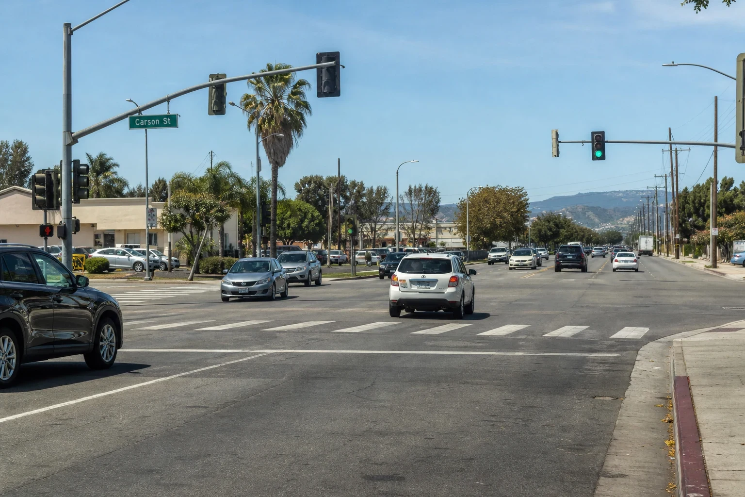 Wide view of a sunny multi-lane intersection on Carson St with traffic lights, palm trees, a crosswalk, everyday cars and low hills in the distance