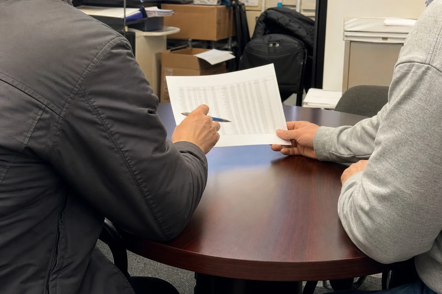 Two people at a small round wood conference table reviewing a printed page of figures, one holding a pen, with boxes and filing cabinets behind