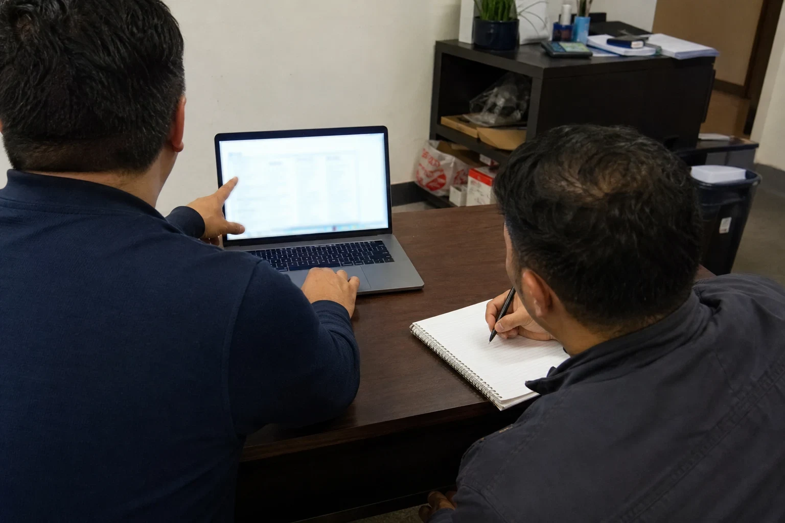 Two men seen from behind at a wooden office desk, one pointing at a laptop screen while the other writes notes in a spiral notebook